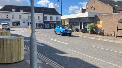 A town centre road facing a junction. There are shops and two cars on the road going in opposite directions. There are some empty wooden planters to the left of the image and bike racks on the right.