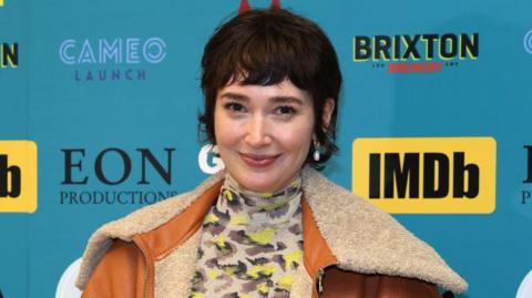 A woman looking at the camera and smiling, she has dark hair and is wearing a brown coat with cream fur. She also has a top with a yellow and brown design which covers her neck. She is standing in front of a board which has sponsor logos including 'IMDb', 'Brixton Brewery', 'Cameo Launch' and 'EON Productions'.