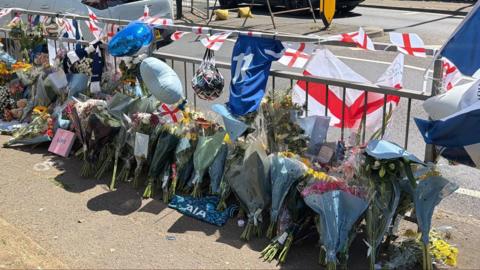 Several flowers, England flags and football merchandise are either standing against or are tied to a silver fence which runs alongside a single carriageway.