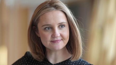 A woman with shoulder-length blonde hair, looking off camera. She is wearing a black and white polka dot top.