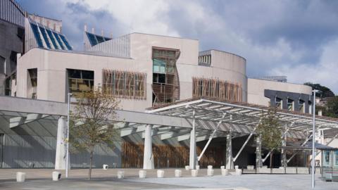 The front of the holyrood building on a cloudy day, with white stone walls wooden slatted windows and small trees.