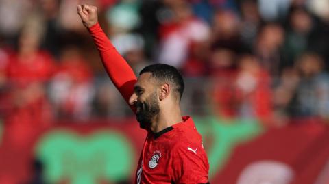 A smiling Mohamed Salah in red Egypt Jersey, visible from shoulders up, is seen looking from right to left and raising his right arm, the one furthest from the camera, high in the air straight up above his head and clenching his fist