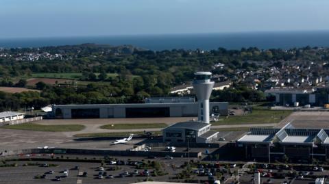 Aerial view of an airport. Cars are parked in a large car park. An air traffic control tower is in the centre. Residential properties can be seen in the distance as well as trees and the sea.