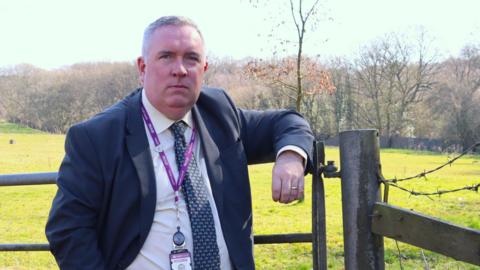 Councillor Adrian Andrew, deputy leader of Walsall Council, stands leaning on the side of a metal gate. He is wearing a suit and tie with a purple lanyard around his neck. A field and trees can be seen behind him.
