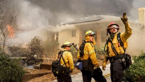 Three Los Angeles County firefighters look away from camera and gesture next to a burning home while trying to protect homes from the Eaton wildfire in Altadena on Wednesday