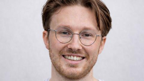 Fergal Mackie smiling at the camera. He has brown hair and is wearing glasses.