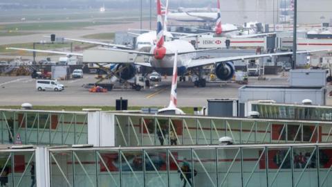 A general view of Heathrow Airport, with glass walkways showing passengers walking and planes on a runway.