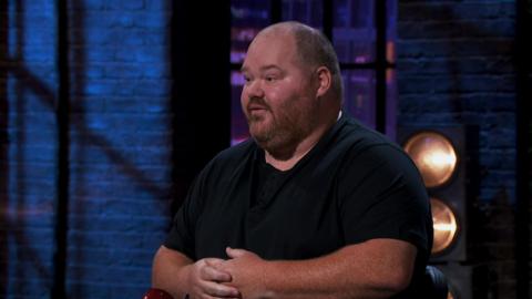 A close up shot of Simon Sansome sitting in front of the dragons pitching his app. He is wearing a black t shirt.