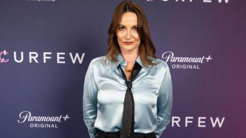 Sarah Parish is standing in front of a purple background in a blue silky blouse and a tie. She has long brown hair and is looking at the camera.