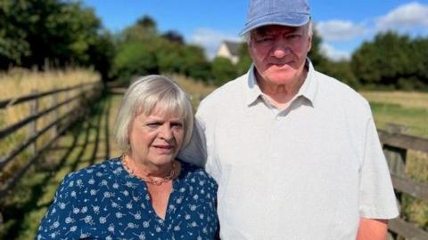 Pat Ferguson and Chair of Preston Parish Council, Councillor Ian Ireland, stand on part of the public footpath called Nuns Walk. Pat Ferguson has blonde hair in a short bob, is wearing a blue shirt and gold necklace. Ian Ireland is wearing a baseball cap and white shirt. Both are looking into the camera with concerned expressions.