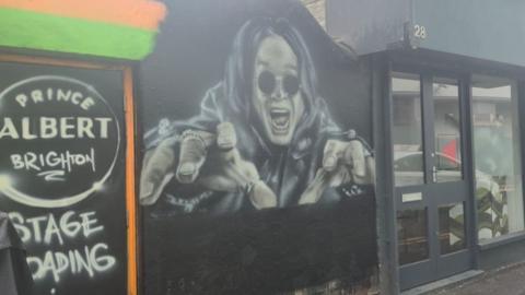 A painting of Ozzy Osbourne with his hands outstretched on the side of a black wall.