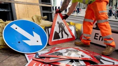 A man in high-vis trousers holding a red roadworks sign among a stash of temporary signage. There are other signs surrounding him, including a blue one-way sign with an arrow