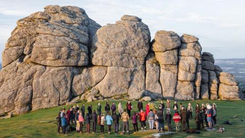 A large group of people are stood in a circle next to part of Haytor - a large set of grey craggy rocks on Dartmoor.