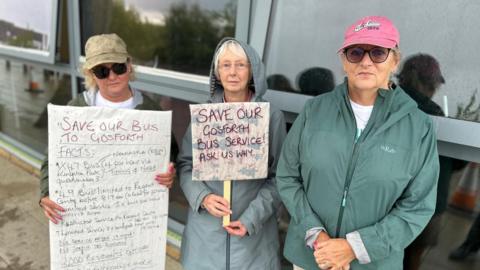 Three people holding signs. One of the signs say: "Save our Gosforth Bus Service! Ask us why"