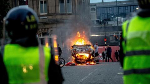 Two police officers in riot gear stand in the foreground of the shot. They face away from the camera towards a burning car in the middle of the road.