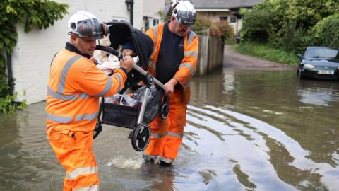 Two men in orange hazard suits and hard hats carry a buggy with a sleeping baby through flood water on a residential street