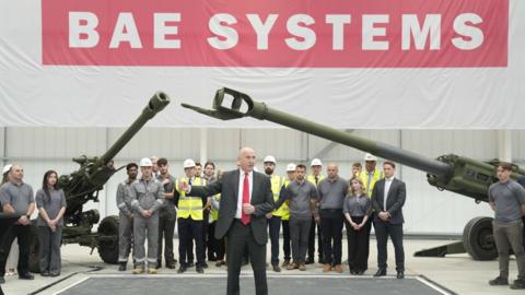 A man in a smart suit and red tie stands delivering a speech in front of a large group of people in hard hats and safety jackets. They all stand under two large artillery pieces.