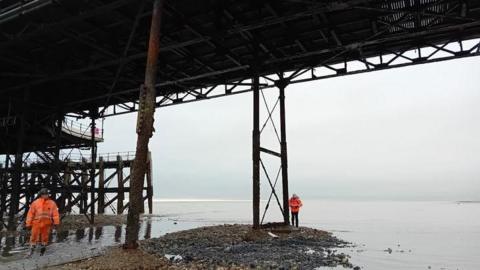 Two contrators in hi-vis stand underneath the pier next to a support and one pile holding up the pier from the sea shore.