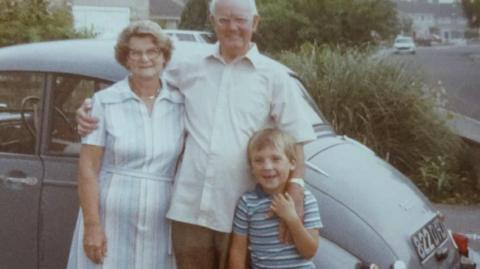 An old photograph of an elderly couple standing in front of a grey classic car with their grandson. The man has his arms around the woman and the child. All of them are smiling. 