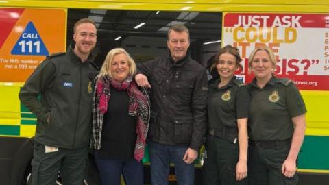 A man and a woman are standing alongside three ambulance crew members in uniform. They are standing by the side of an ambulance with West Midlands Ambulance Service livery.