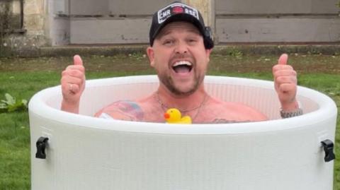 Image of Harry Beattie in a round ice bath, smiling at the camera with his thumbs up, all he is wearing is a black cap.