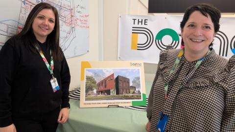 Kelly Prince on the left and Ellen Birchenough on the right, with a CGI image of the completed building on a board in between them both. Prince has long dark hair and wears a black top and trousers - Birchenough has a black and white checked top and short dark hair.