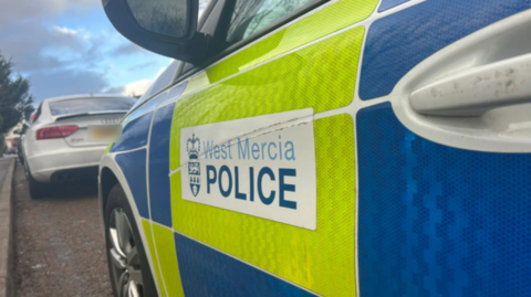 The side of a white police car with yellow and blue squares on the side.
