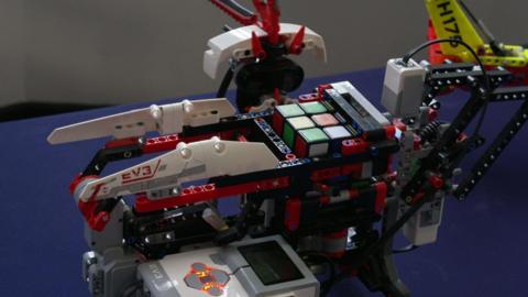 A white and red robot holding a rubik's cube. It is stood on a blue table. 