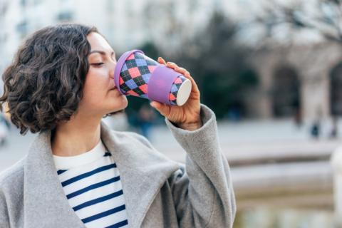 Young woman with dark hair in a short bob wearing a grey wool coat over a blue and white stripped top drinks from a reusable cup