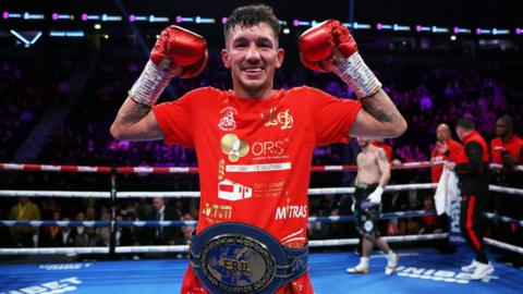 Liam Davies is posing in a blue boxing ring.  He is smiling and is wearing red boxing gloves with his arms raised in the air. He is wearing a red t-shirt and a large blue boxing trophy belt.
