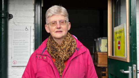 A woman, with grey hair and wearing a pink coat, and leopard print scarf , stood in a doorway beneath a sign saying "Welcome to Topsham Museum"