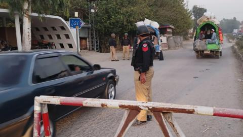 A police officer in a black helmet, black shirt and beige trousers stands guard at a checkpoint along a dusty highway in the Khyber Pakhtunkhwa province