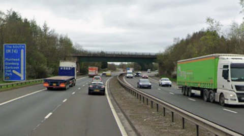 Cars going both ways on a dual carriageway with a bridge overhead. Trees border the road and it is a cloudy day