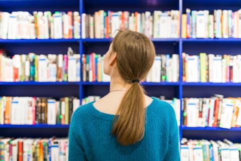 "Rear view of a person with long brown hair in a ponytail, looking at bookshelves filled with books on vibrant blue shelves."