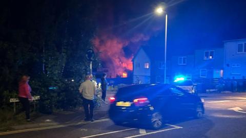 Four people stand beside a car at night with a fire raging in the background
