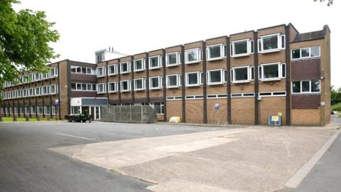 The brick buidling has three floors and two blocks interlinked. There are many uniformed white windows along each floor. The block on the right is boarded up on the first floor. A concrete car park sits in front of the building.