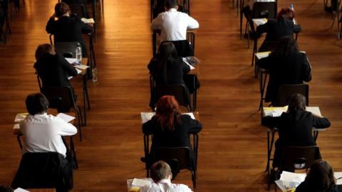 Pupils at a secondary school sit at tables while taking exams in a hall. All the children have their backs turned to the camera and papers and pencil cases strewn across their desks.