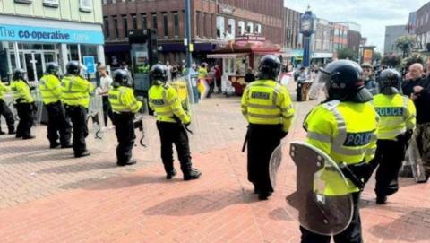 Police officers in a line, wearing riot gear and holding shields, stand near the Blue Clock in Hanley


