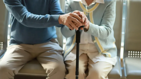 A mid shot of two elderly people. They are both sitting on chairs and holding hands and a walking stick. The person on the left is wearing a blue jumper and light trousers. The person on the right is wearing a light blue cardigan and light trousers.