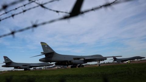 Three large planes are stationed on a runway. In the shot is also barbed wire.