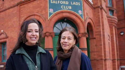 Laura Slingsby (left) chief executive at Salford Lads and Girls Club and Claudia Kenyatta of Historic England pose for a picture outside Salford Lads and Girls Club. Laura has long dark brown hair and is wearing a black polo neck under a green corduroy dress and black coat.Claudia Kenyatta has bobbed brown hair and wears a brown scarf and navy blue top with black trousers. They are both smiling.