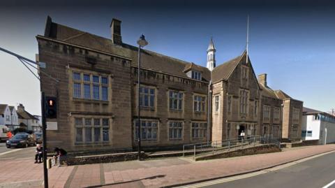 A Google Street View screenshot of Carlisle Magistrates' Court which is a large, brown brick, corner building with many windows.