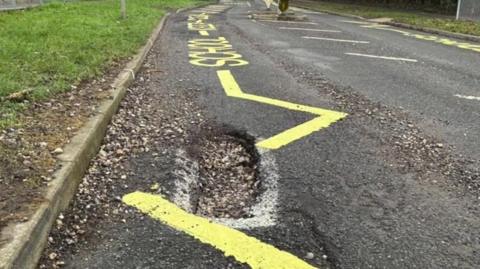 A road with a large pothole in it close to the curb. Some freshly painted yellow school markings are zig-zagging the road and go directly over the pothole.