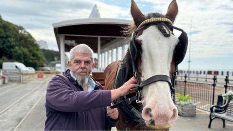 Peter Cannon holding the strap of a brown and white horse standing in front of a horse tram.