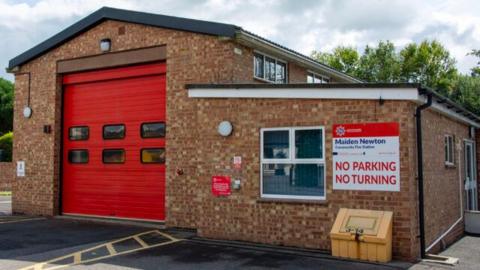 A typical fire station building with a large red garage-style door for vehicles. A sign to the right of the building reads "Maiden Newton Community Fire Station". Underneath that the sign reads "NO PARKING" and "NO TURNING". 