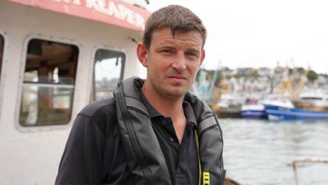 Tom Parker is staring straight into the camera. He is standing on his fishing boat in the harbour, and wearing a black T-shirt and a black plastic life jacket. Behind him is Brixham harbour, with fishing boats in the harbour. 