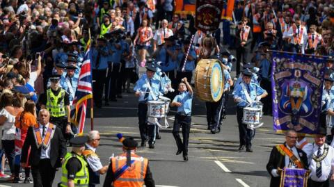 A parade of people wearing dressed in orange and blue uniforms, wearing caps and carrying drums and other instruments march in rows in Glasgow in 2019.