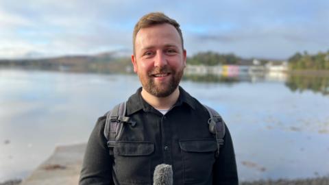 Jamie McDonald holds a microphone, behind him is a loch on the Isle of Skye 