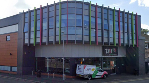 A van with 'Inspire Youth Zone' on the side is parked in front of a large building which has colourful vertical columns on the windows on the first floor and a colourful sign saying 'Inspire Youth Zone' above the door.