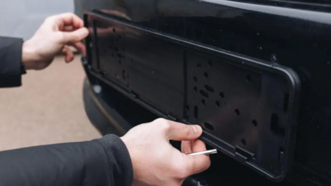 Two hands, one holding a metal tool, are pictured touching the space where a number plate should go at the back of a black car